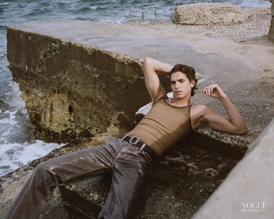 Male model reclining on coastal stone steps wearing a tan knit tank top and dark glossy trousers by the sea.