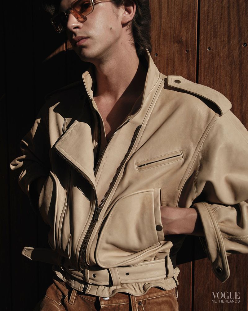 Model wearing an oversized beige leather jacket, amber tinted sunglasses, and brown trousers against vertical wood paneling.