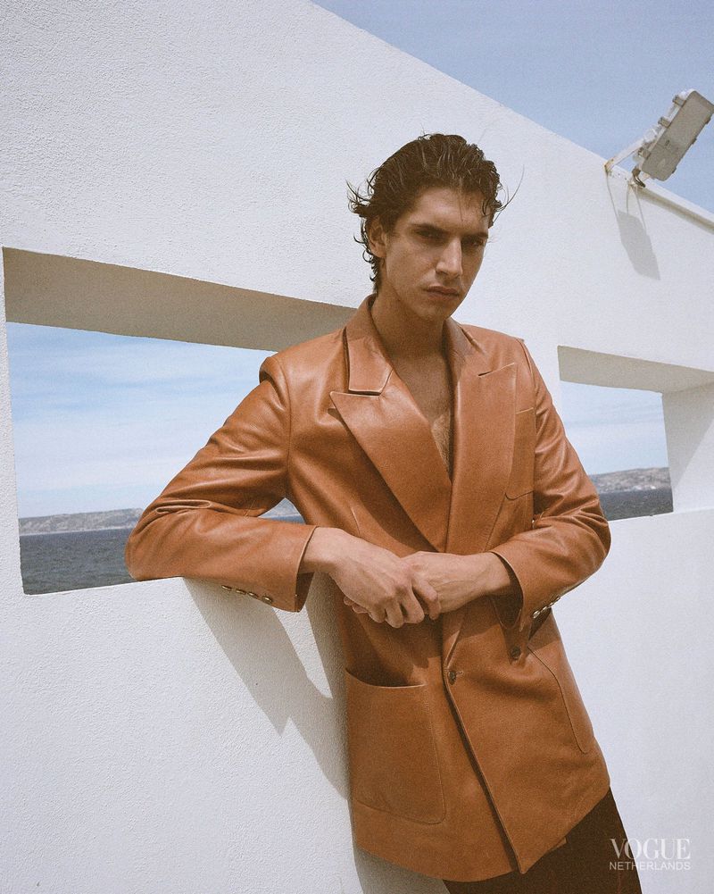Male model wearing a caramel brown leather blazer leans against a white architectural wall overlooking the sea.