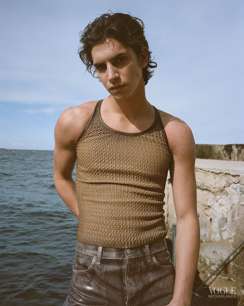 A young man wearing a textured olive tank top and grey denim jeans stands outdoors beside the ocean against a stone pier.