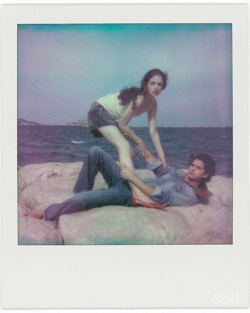Polaroid style photo of a man reclining on coastal rocks and a woman standing over him near the water.