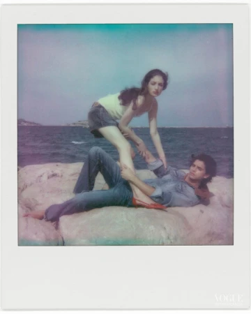 Polaroid style photo of a man reclining on coastal rocks and a woman standing over him near the water.