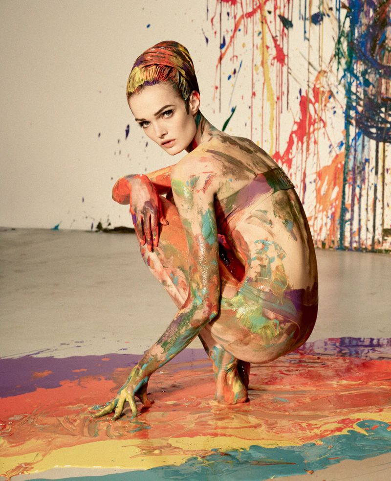 Model Lulu Tenney crouching covered in multicolored paint on a studio floor with paint splatters.