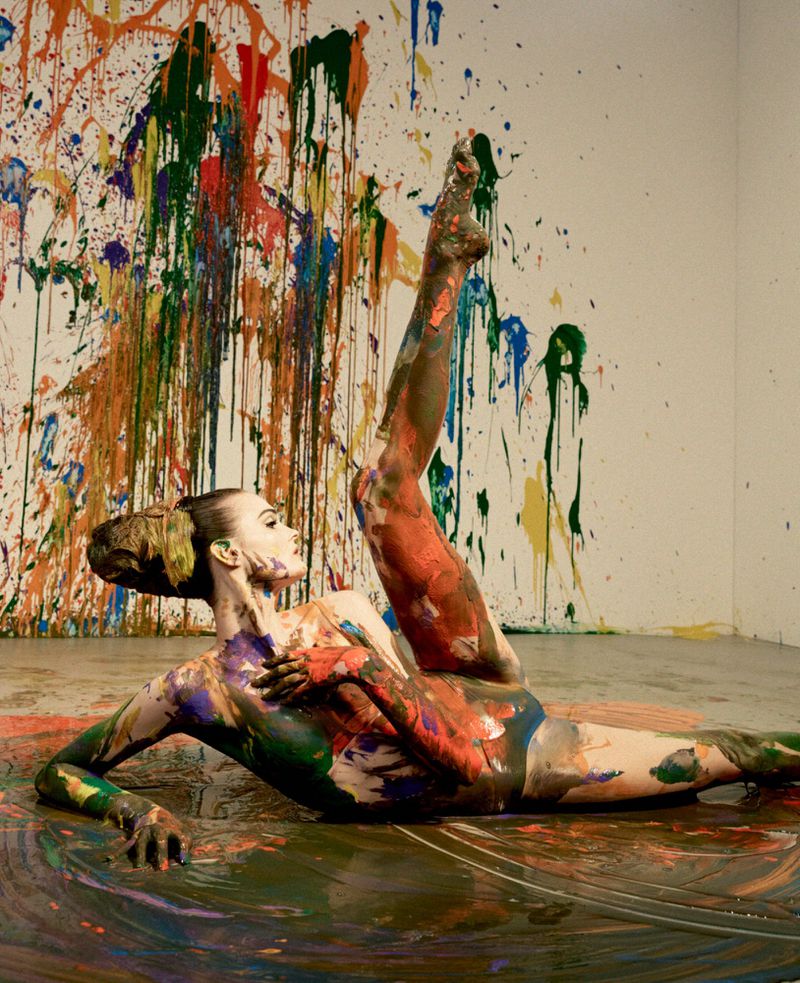 Model covered in multicolored paint posing on a paint-slicked floor with one leg extended upward against a paint-splattered wall.