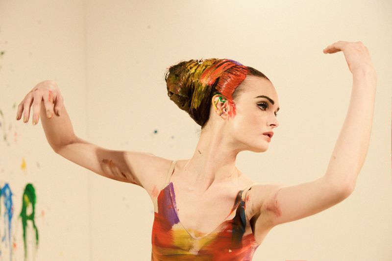 Model Lulu Tenney poses with raised arms, wearing vibrant colorful paint across her styled hair and bodice in a studio setting.
