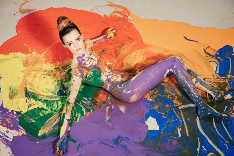 Model Lulu Tenney reclined and covered in vibrant poured acrylic paint across a colorful floor.
