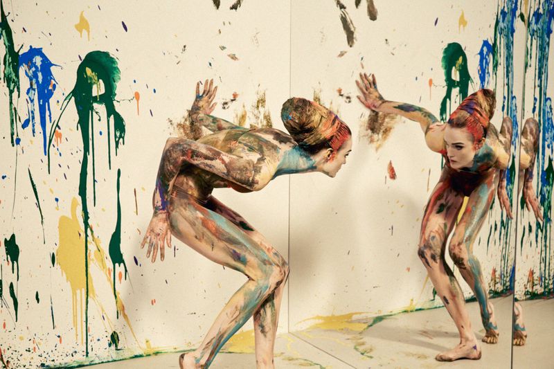 Model Lulu Tenney covered in multicolored body paint poses in front of a mirror and paint-splattered white walls.