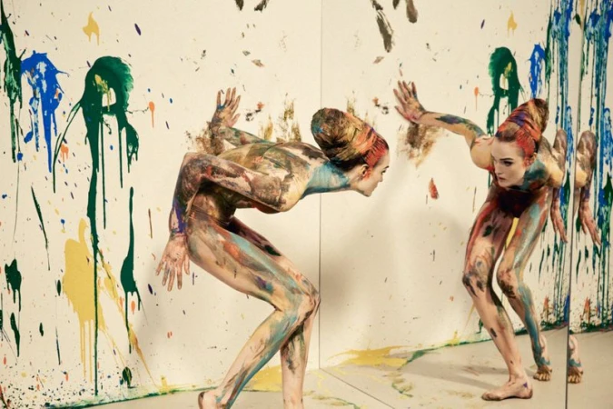 Model Lulu Tenney covered in multicolored body paint poses in front of a mirror and paint-splattered white walls.
