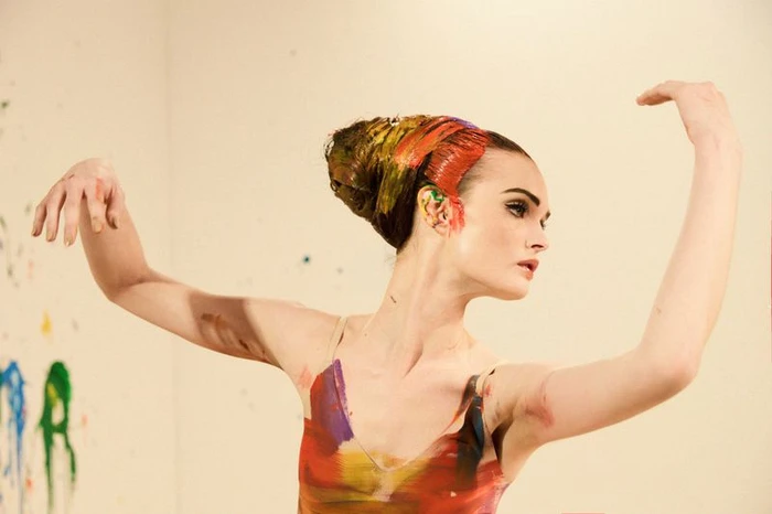 Model Lulu Tenney poses with raised arms, wearing vibrant colorful paint across her styled hair and bodice in a studio setting.
