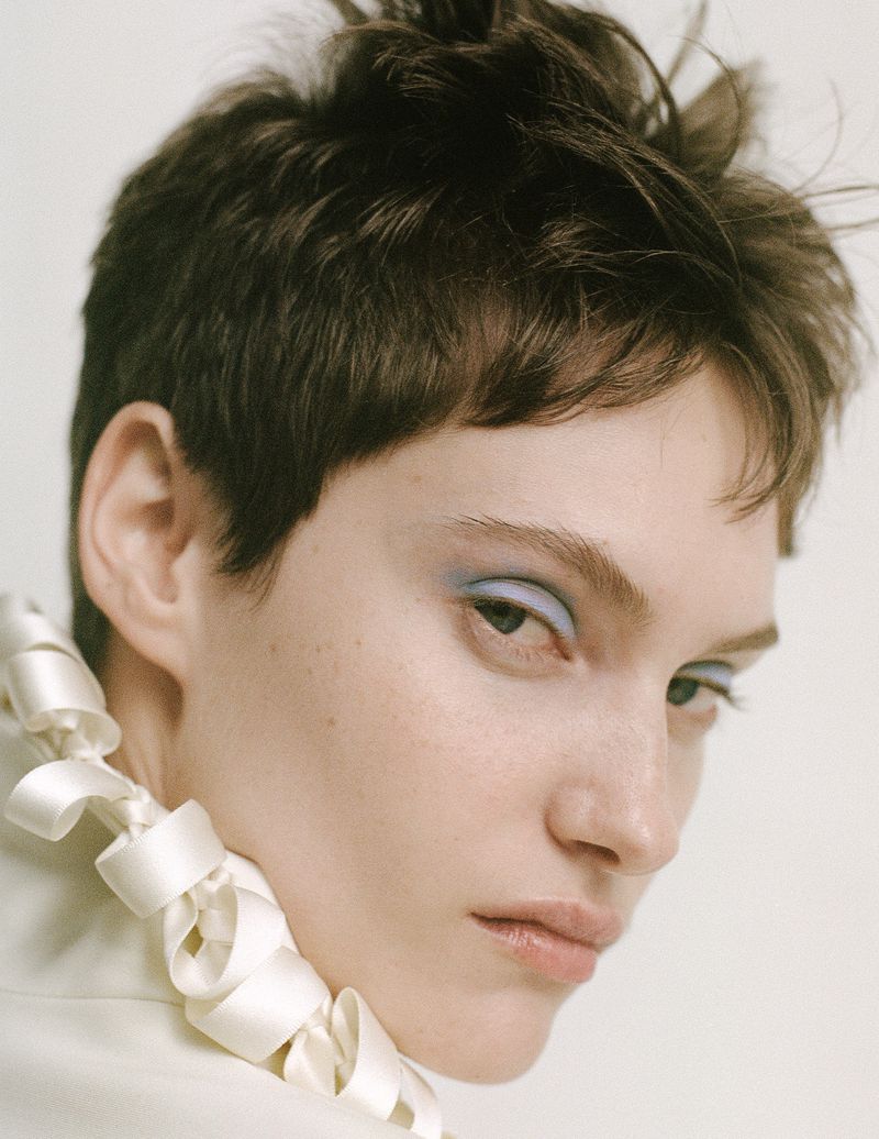 Close-up portrait of model Greta Hofer with short brunette hair, soft blue eyeshadow, and a coiled white ribbon collar against a light backdrop.