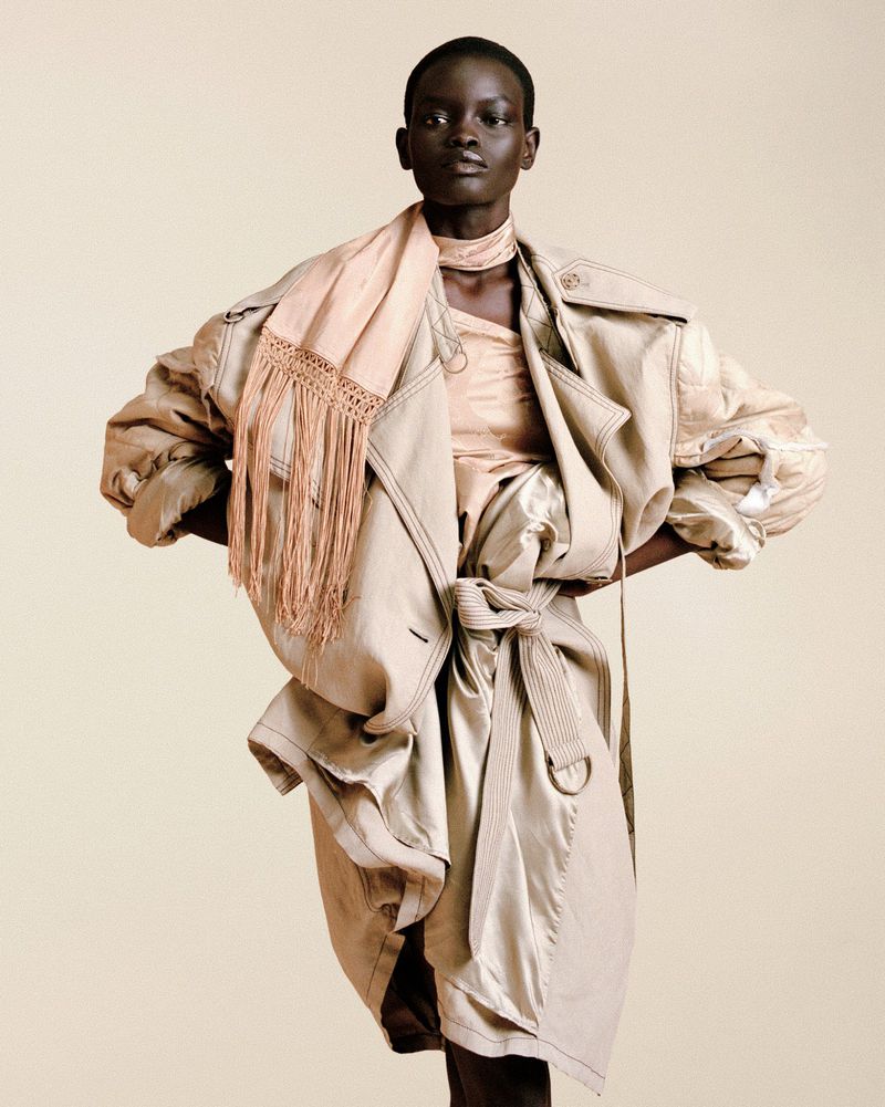 Model wearing an oversized deconstructed beige trench coat with a peach fringed scarf against a warm neutral studio backdrop.