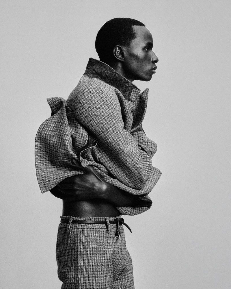 Black and white profile portrait of a Black man wearing a textured houndstooth jacket and trousers against a pale studio backdrop.