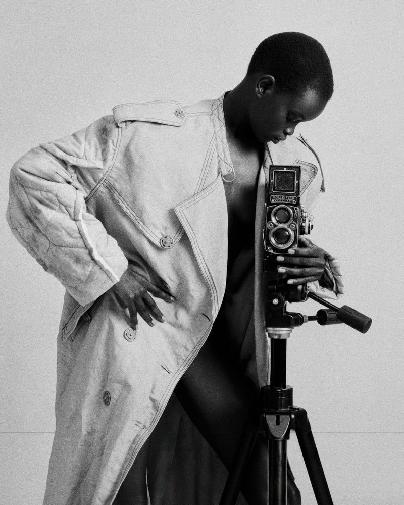 Model wearing an open trench coat poses with a vintage medium format camera mounted on a tripod in black and white.