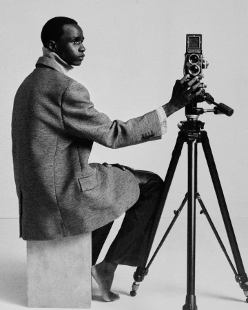 Man in a tailored blazer seated barefoot on a studio block adjusting a twin-lens reflex camera on a tripod against a white backdrop.