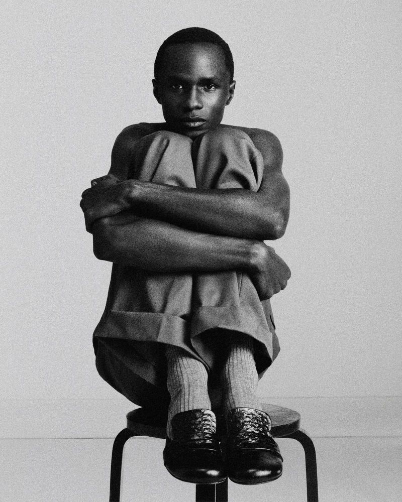 Black and white portrait of a shirtless man seated on a stool hugging his knees to his chest against a studio backdrop.