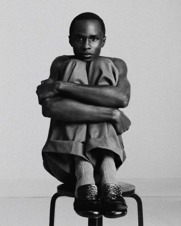 Black and white portrait of a shirtless man seated on a stool hugging his knees to his chest against a studio backdrop.