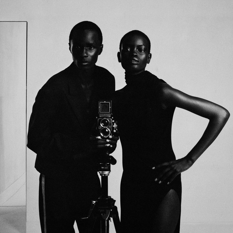 Black and white studio portrait of a man and woman in black standing beside a medium format camera on a tripod.