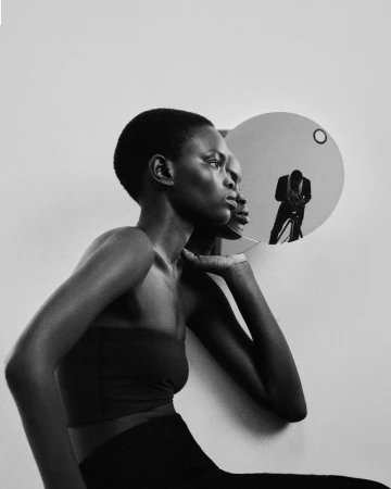 Black and white portrait of model Athiec Geng in a strapless top beside a round wall mirror reflecting the photographer.
