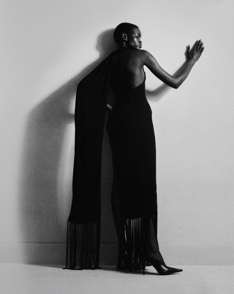 Model Athiec Geng poses in a backless black fringed gown and pointed stiletto heels against a studio wall with a cast shadow.