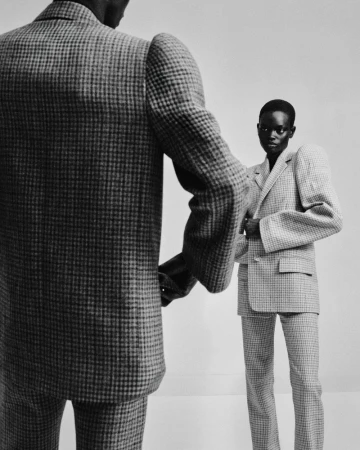 Model wearing a structured houndstooth suit facing the camera behind another model in a matching tailored suit in a studio setting.