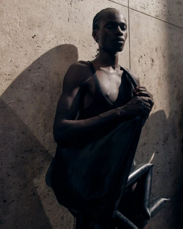 Model in a black knit top holding an oversized black leather bag against a textured stone wall in direct sunlight.