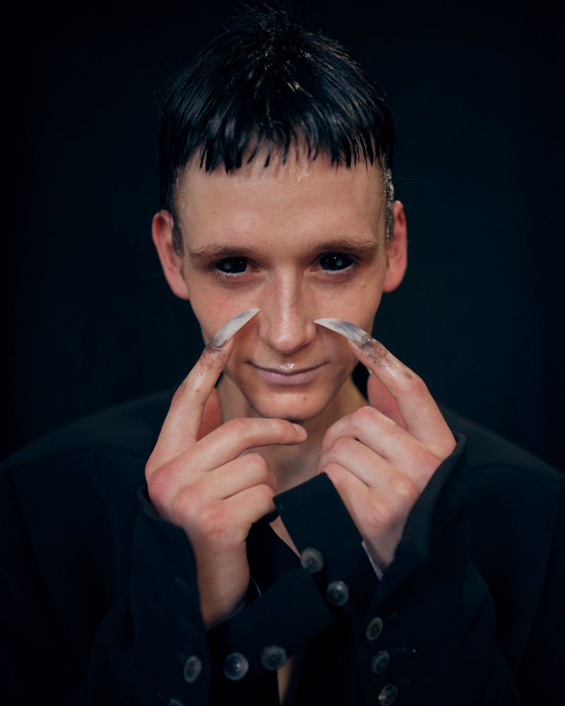 Close-up portrait of a model with black contact lenses and long pointed fingernails wearing a black tailored jacket against a black backdrop.