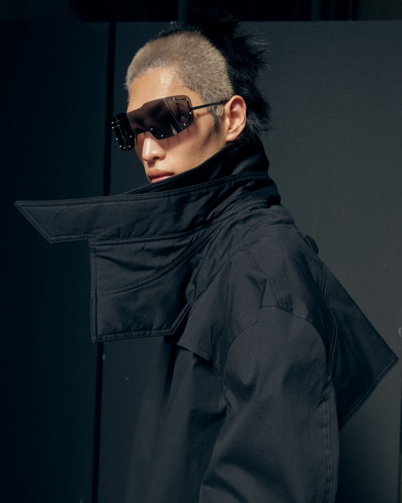 Model wearing futuristic shield sunglasses and a structured high-collar black jacket looking over his shoulder in a dark studio.