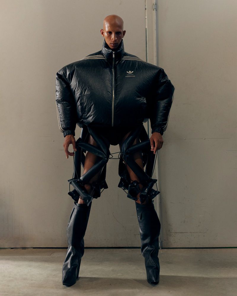 Model wearing an exaggerated black puffer jacket with angular shoulders, sculptural leg cage harnesses, and tall black leather boots.