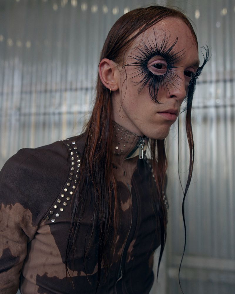 Model backstage at Rick Owens wearing exaggerated radial feather false eyelashes and a studded high-collar jacket.