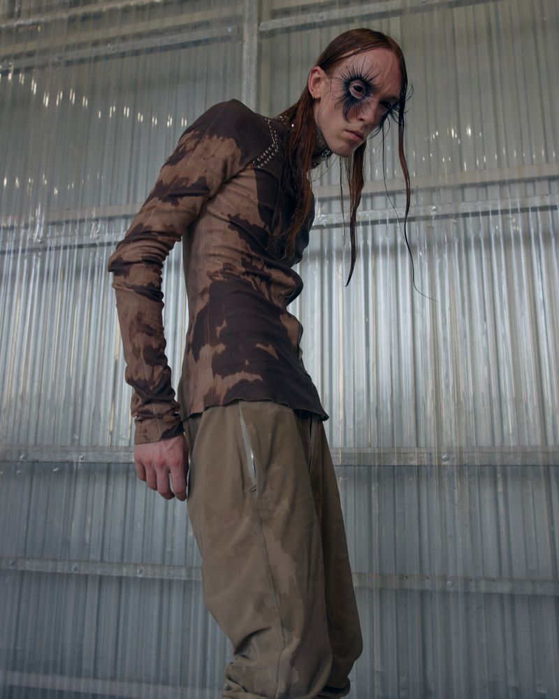 Model with dramatic dark eye makeup posing in a brown patterned long-sleeve shirt and khaki trousers against corrugated panels.