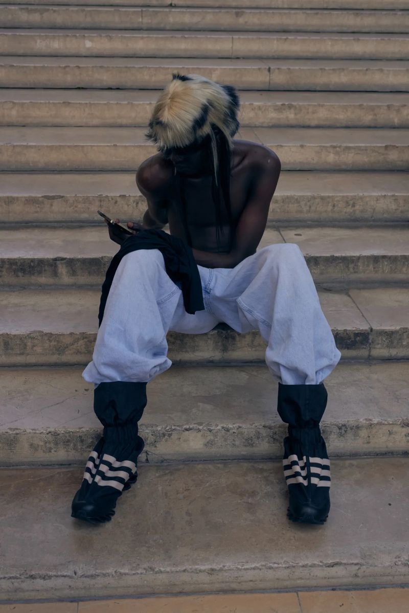 Shirtless model with feathered hair seated on outdoor stone steps looking at a phone in loose trousers and striped boots.