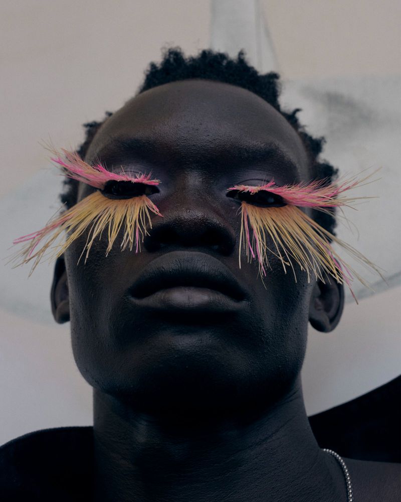Close-up portrait of a Black man wearing long feathered false eyelashes in vibrant pink and yellow hues against a soft backdrop.