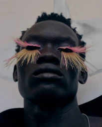 Close-up portrait of a Black man wearing long feathered false eyelashes in vibrant pink and yellow hues against a soft backdrop.