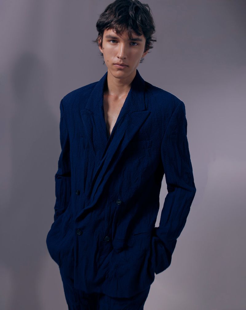 A male model wearing an oversized navy blue double-breasted suit jacket with hands in pockets against a plain grey studio backdrop.