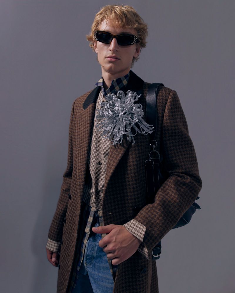 Model wearing dark sunglasses, brown houndstooth coat with a silver floral brooch, and checkered shirt against a grey backdrop.