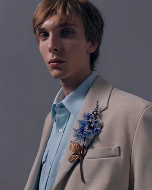 Close-up portrait of a male model wearing a light beige blazer adorned with a blue beaded floral brooch and a pale blue shirt.