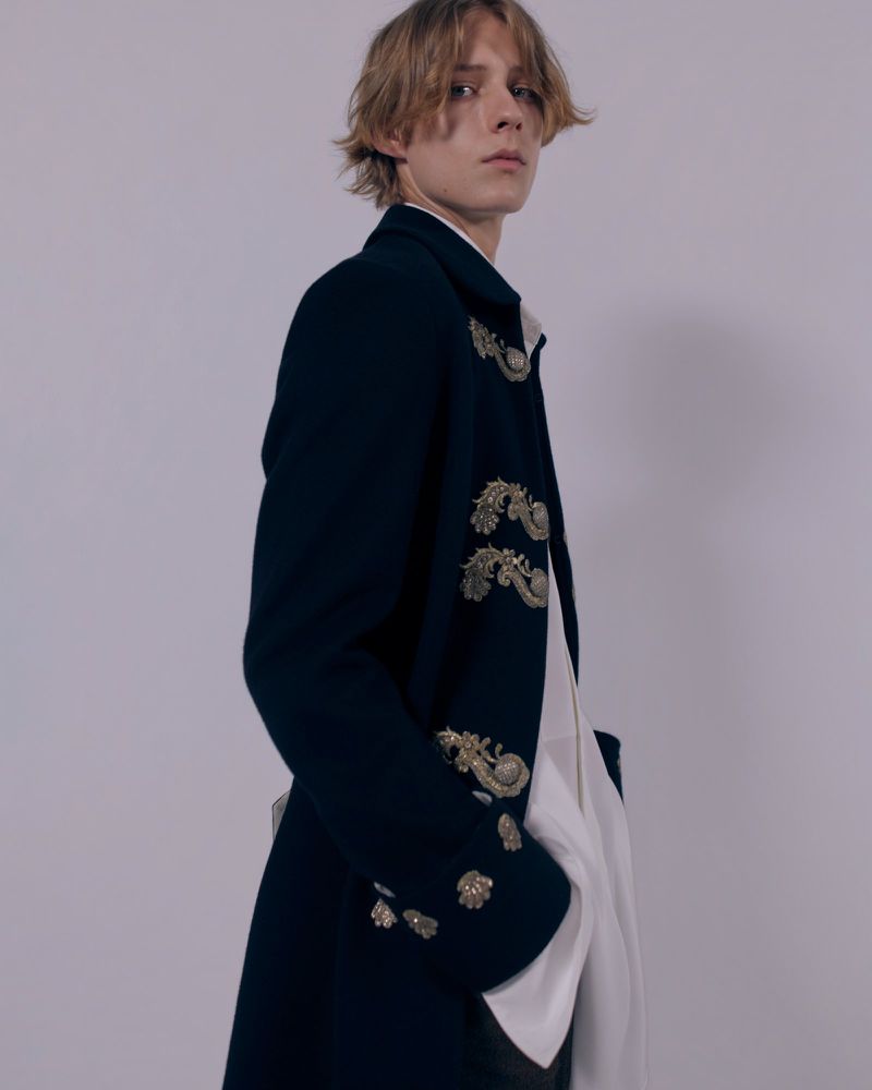 Model wearing an embroidered dark navy coat with metallic frogging over a white shirt against a neutral studio backdrop.
