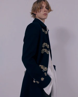 Model wearing an embroidered dark navy coat with metallic frogging over a white shirt against a neutral studio backdrop.