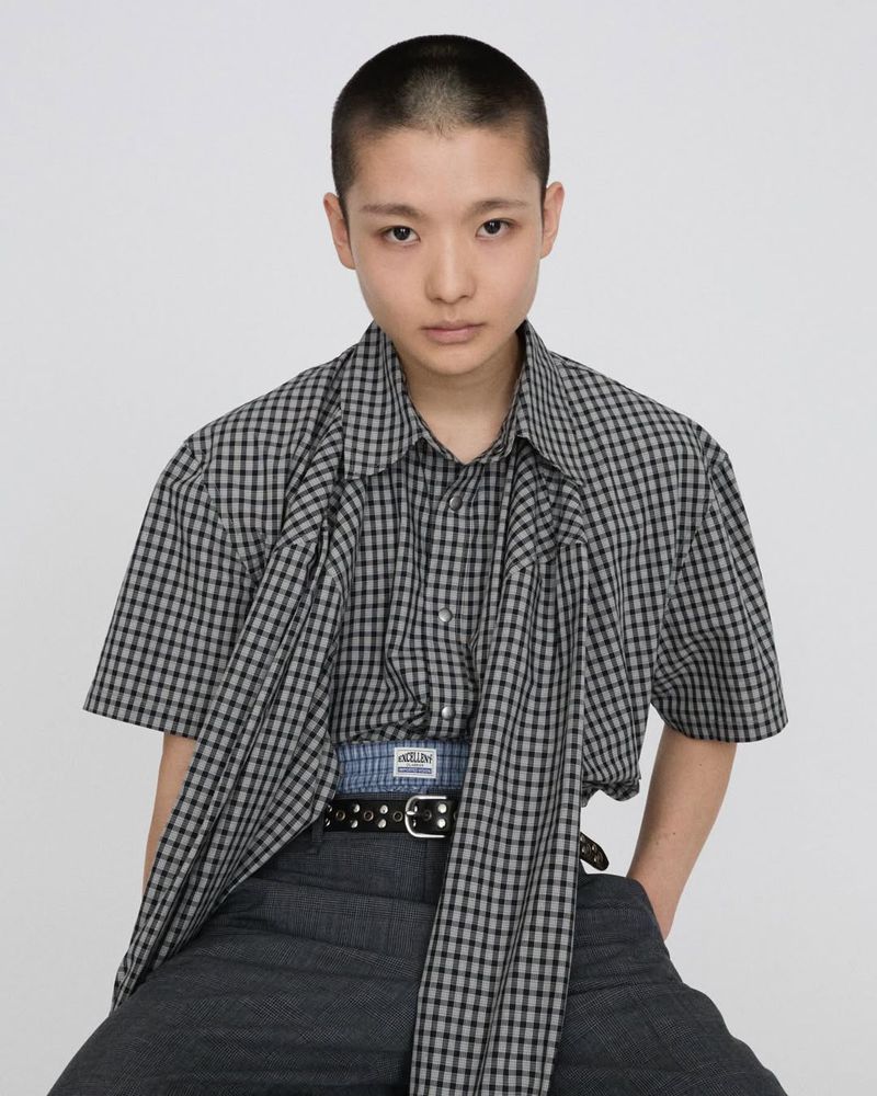 Portrait of a young adult in a checkered shirt and grey pants against a white backdrop, demonstrating modern casual fashion.