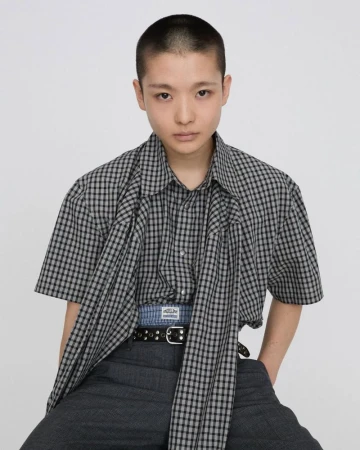 Portrait of a young adult in a checkered shirt and grey pants against a white backdrop, demonstrating modern casual fashion.