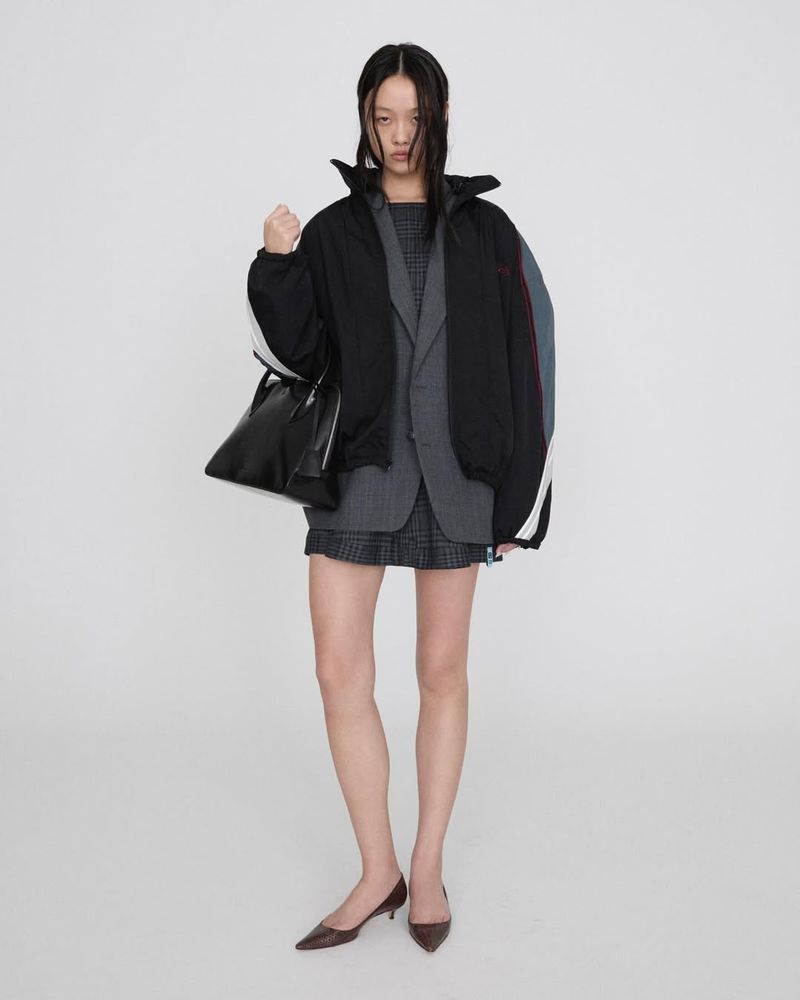 Studio shot of a stylish adult woman in a blazer, skirt, jacket, and heels, carrying a black purse against a plain white backdrop.