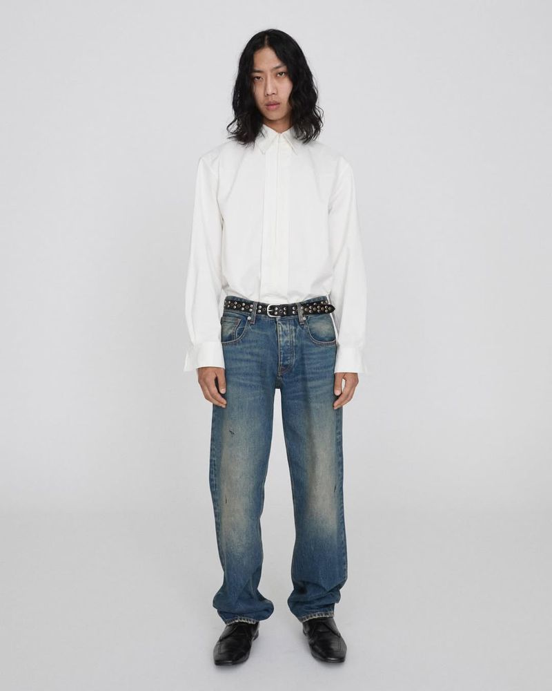 Full shot of an adult with long dark hair, wearing a white button-down shirt, jeans, black studded belt, and black leather shoes.
