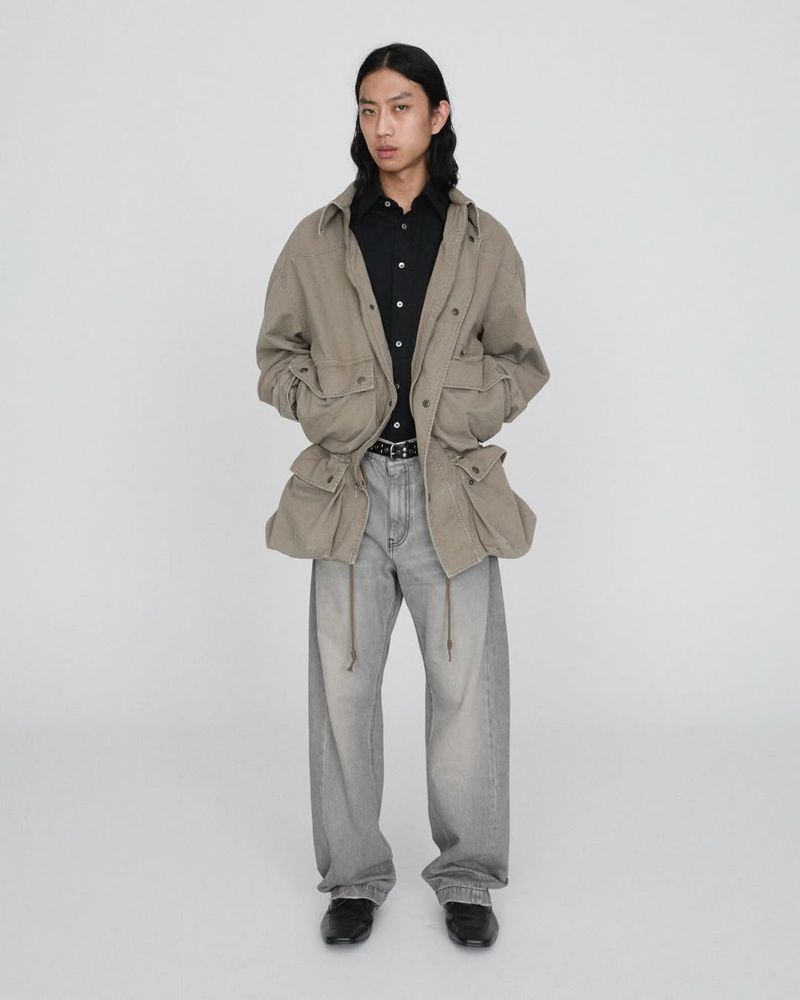 Full-length shot of an Asian man wearing a khaki jacket, black button-down shirt, grey jeans, and black shoes posing in a white photo studio.