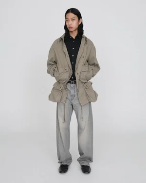 An East Asian male model stands in a khaki multi-pocket jacket, black shirt, and faded grey wide-leg jeans on a grey studio backdrop.