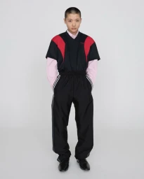 Model standing against a plain white background wearing a layered black top with red panels over a light pink collared shirt and black trousers.