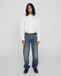 Model standing against a white backdrop wearing a white button-down shirt, straight-leg blue denim jeans, and a black studded belt.