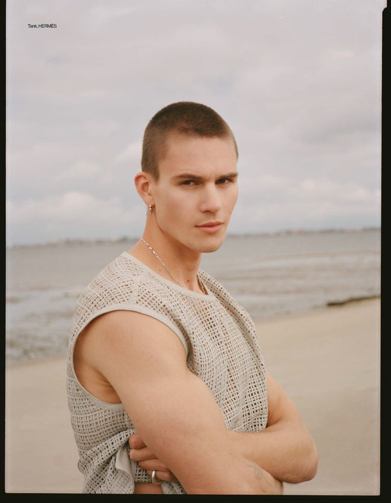Model Samuel Watson posing with arms crossed on an overcast sandy beach wearing an open-knit sleeveless top.