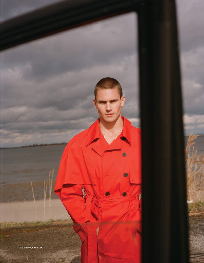 Model Samuel Watson wearing a belted bright red trench coat, framed through a window reflection against a cloudy coastal landscape.