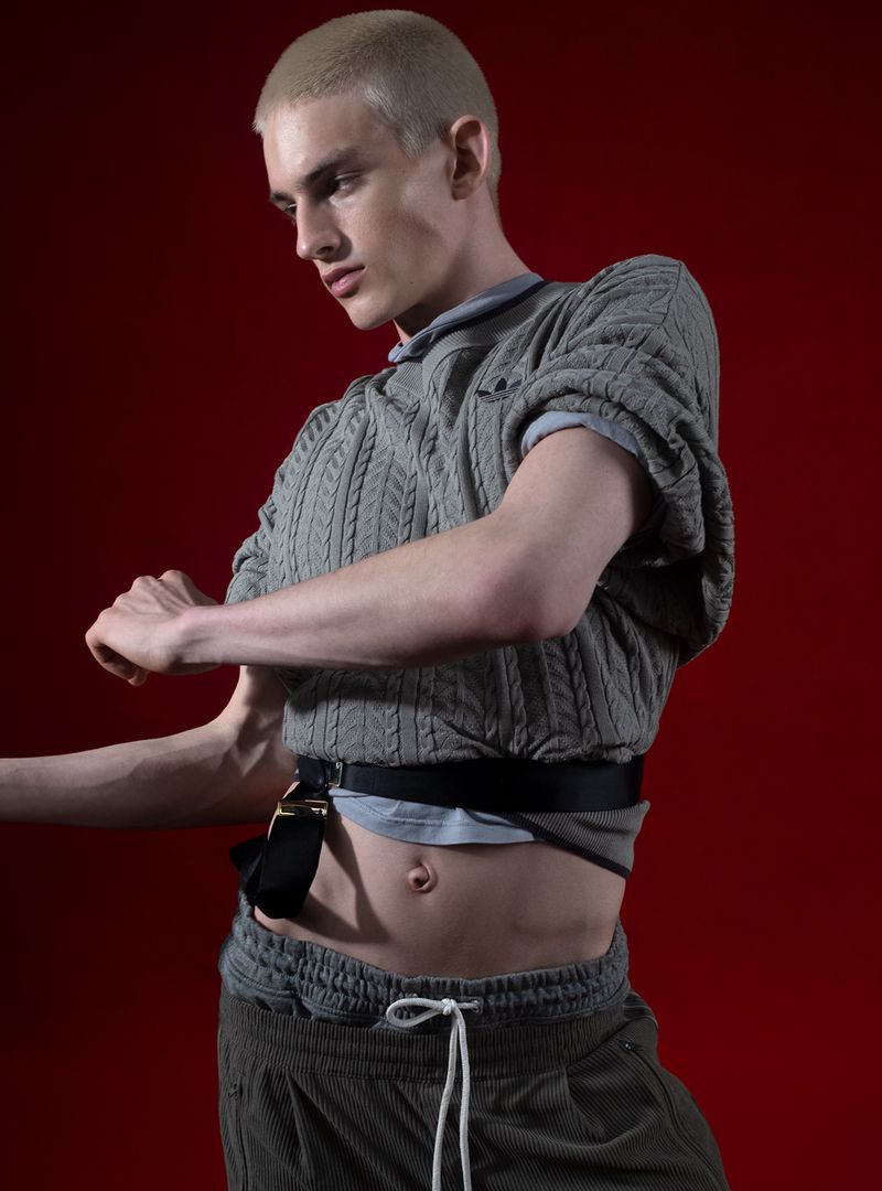 Model with a blonde buzzcut poses in a cropped grey cable-knit sweater and olive trackpants against a deep red backdrop.