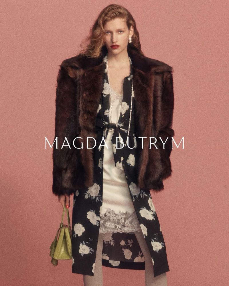 Model Apolline Rocco Fohrer in a brown fur jacket, floral robe, and slip dress holding a green handbag against a pink backdrop.